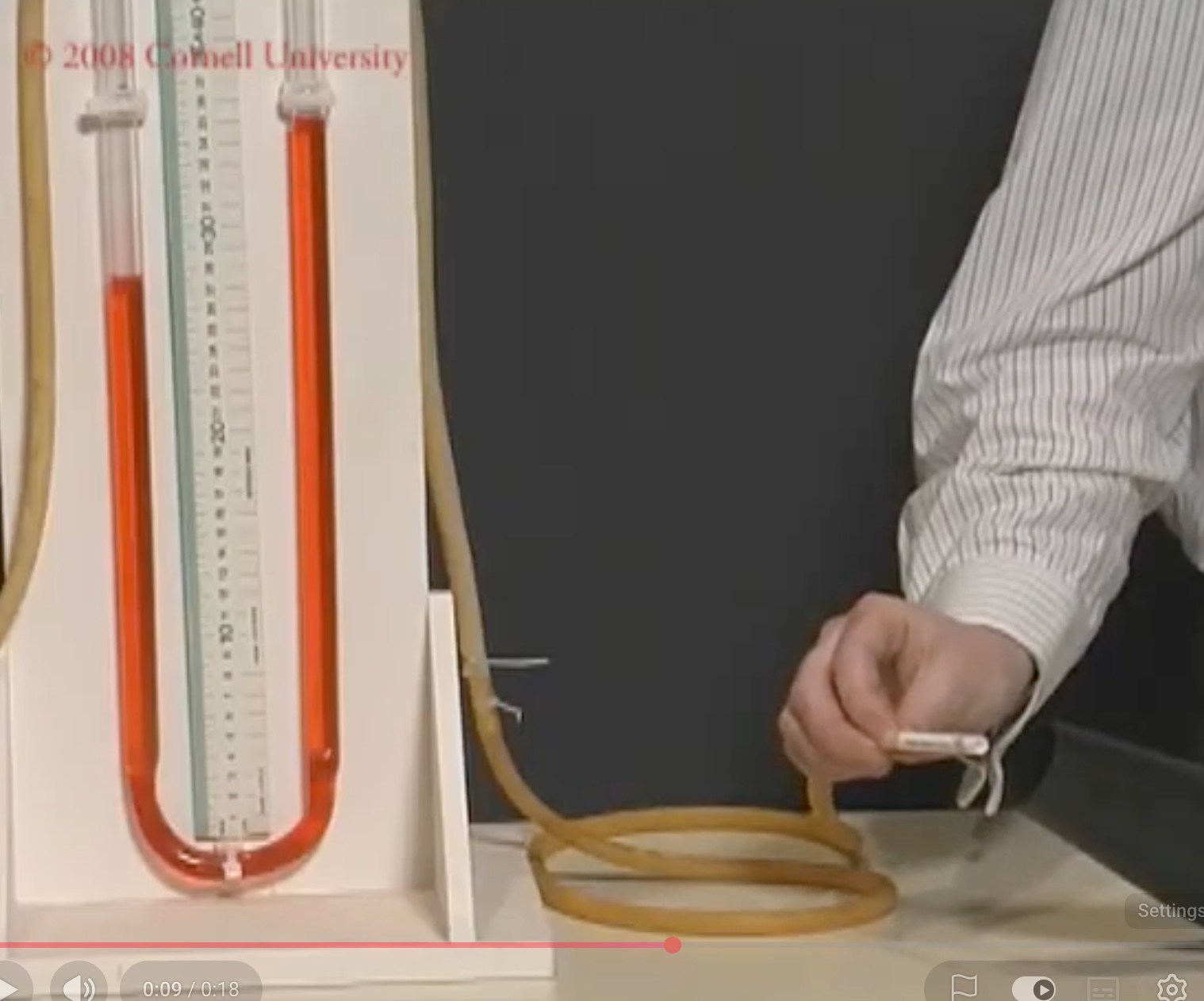 A hand holding a small tube connected by rubber tubing to one arm of a U-tube containing red liquid, whose two levels differ.